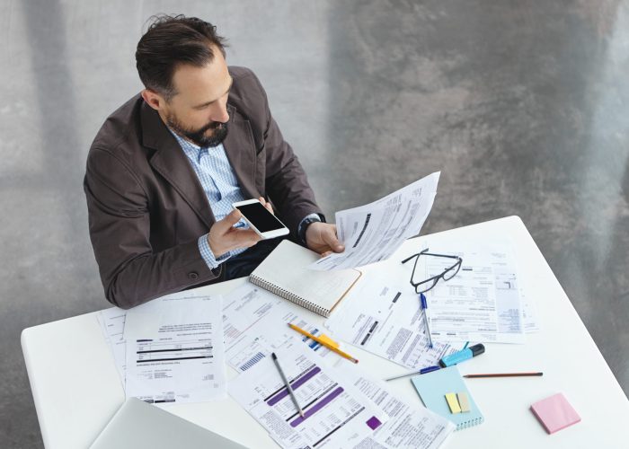 Portrait of busy male office worker makes voice call, discuss documents with partner, compare result, try to predict wastes for next month. Head executive officer holds balance sheet and mobile phone